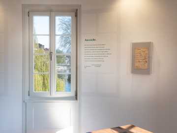 Ein Raum in der Ausstellung. An der Wand kann man Hölderlins Gedicht ›Aussicht‹ lesen. Daneben hängt in einem Rahmen. Darin kann man das Papier sehen, auf das Hölderlin das Gedicht geschrieben hat.