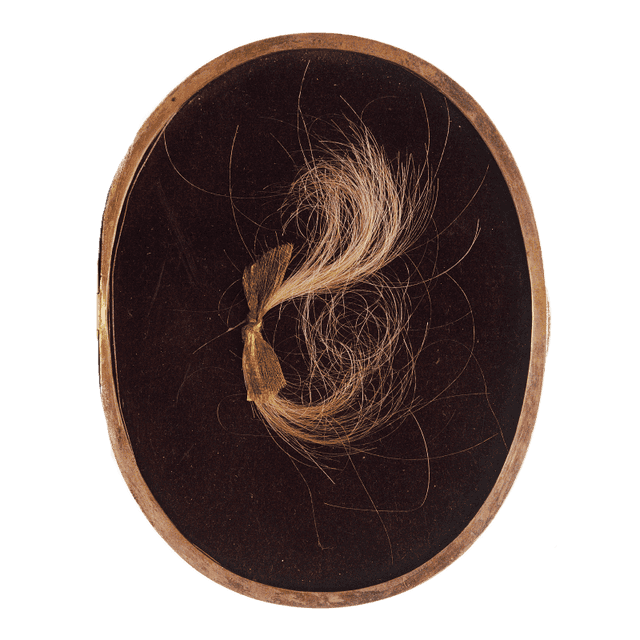 Ein Medaillon mit einer hellbraunen Haarsträhne. Sie wird von einer goldenen Schleife zusammengehalten.