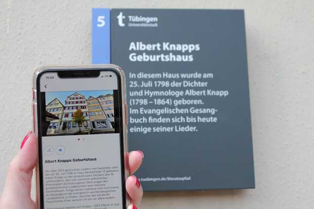 Ein Schild mit der Aufschrift »Albert Knapps Geburtshaus«. Eine Person steht davor und hält ein Smartphone mit einem Hör- und Lesetext dazu in der Hand.