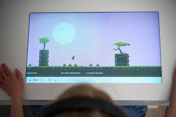 Auf einem farbigen Bildschirm ist ein Spiel mit einer springenden Figur zu sehen. Eine junge Besucherin sitzt vor dem Bildschirm. Sie hat einen Kopfhörer auf.