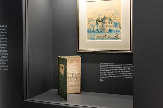 Eine Vitrine mit einem grünen, alten Buch. Dahinter hängt ein Aquarell-Bild vom Neckar am Hölderlinturm.