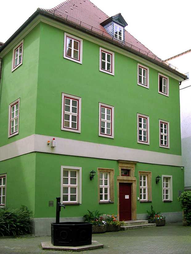Ein Haus mit drei Stockwerken. Das Haus hat eine kräftige, grüne Farbe. Vor dem Haus steht ein Brunnen.