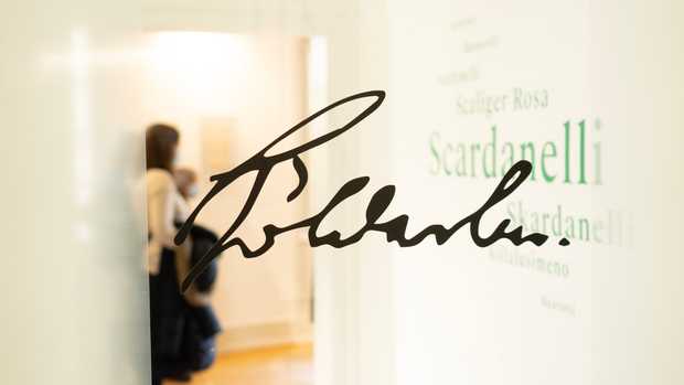 Hölderlins Unterschrift auf einer Glasscheibe hinter der eine Gruppe von Besuchern unscharf zu erkennen ist.